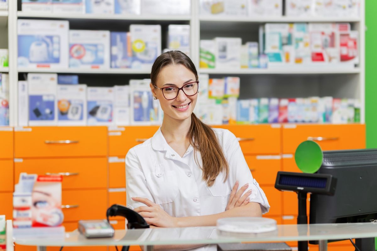 A youthful pleasant dark-haired girl with glasses,dressed in a medical overall,greets visitors at the cash desk in a new pharmacy.